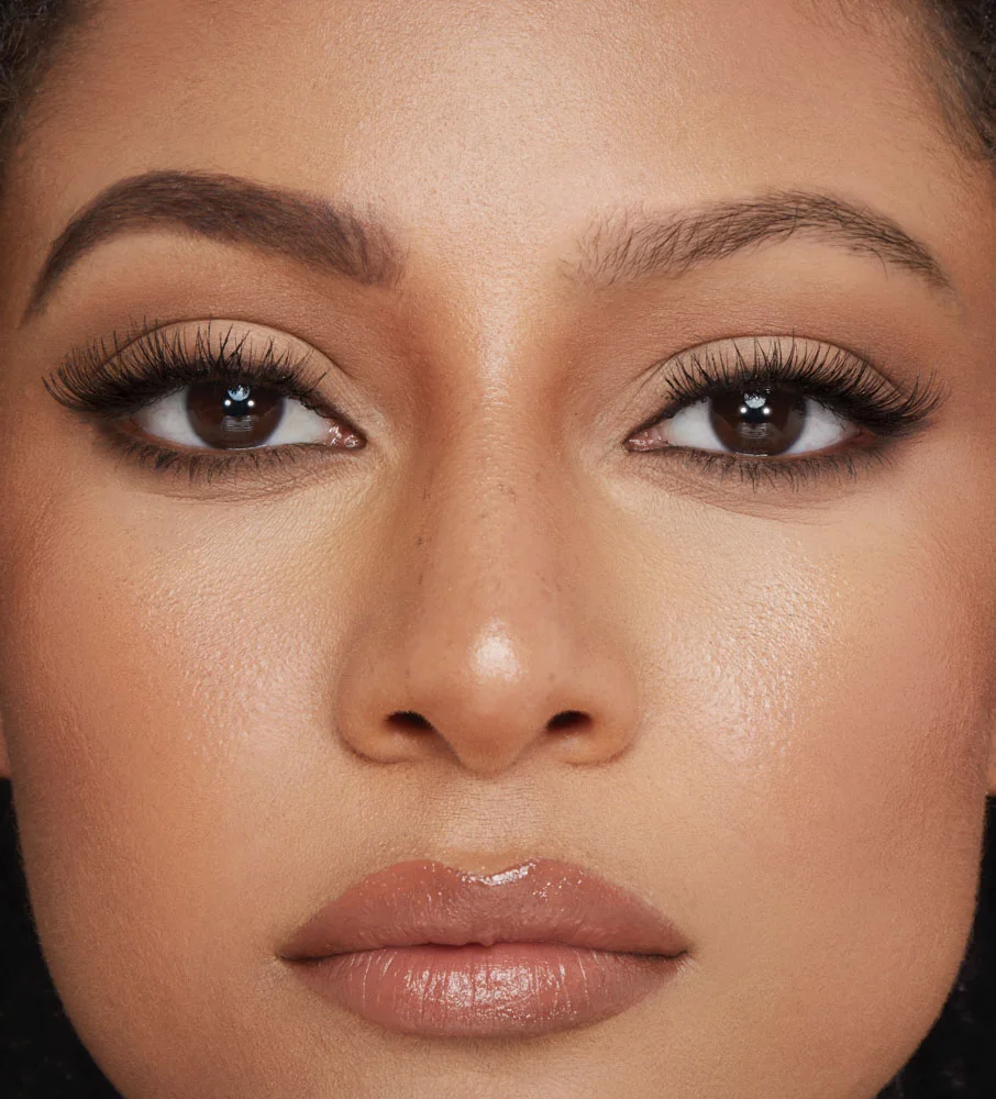 Close-up of a medium-tone model with brown eyes with one eyebrow completely bare and the other filled and fluffed, looking lush and full while also wearing black kohl pencil and nude lip gloss.